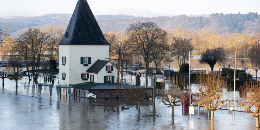 Älteres Gebäude mit Turm am Fluss, umgeben von Bäumen, steht im Hochwasser.