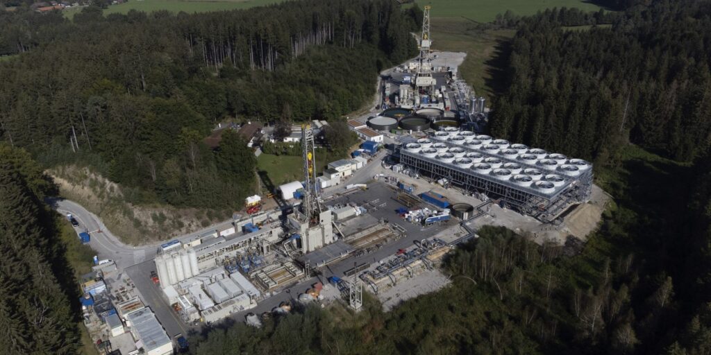 Luftaufnahme des Geothermie-Standorts Geretsried mit zwei Bohrtürmen, Containern und einer Kühleranlage, umgeben von Wald und Feldern