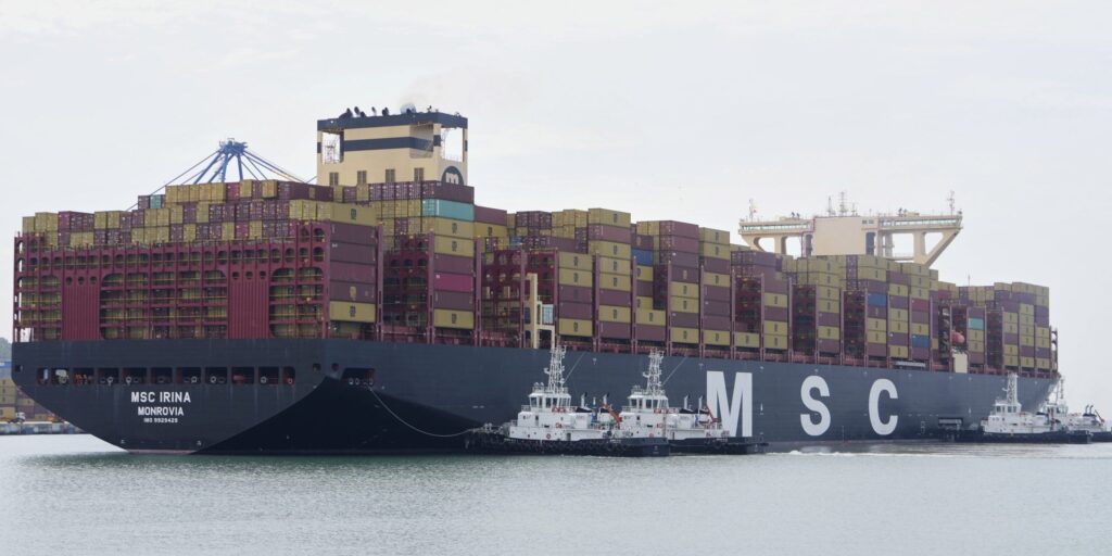 Riesiges Containerschiff MSC Irina mit hohen Containerstapeln in seitlicher Ansicht. Mehrere Schlepper liegen neben dem schwarzen Schiffsrumpf mit großem MSC-Schriftzug.