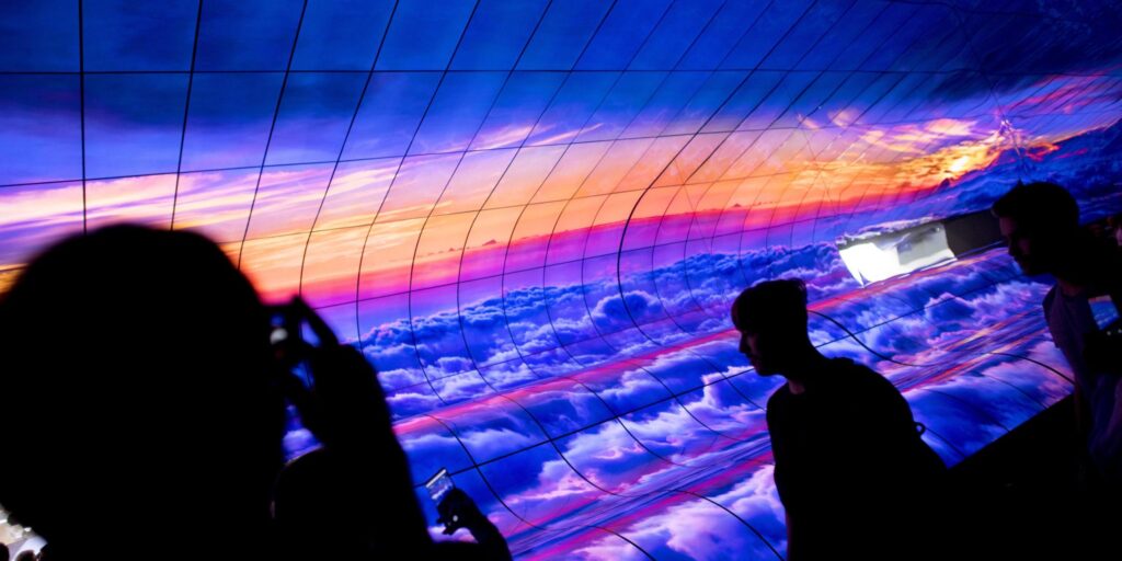 Besucher vor einer gebogenen Displayinstallation mit blauem Himmel, Wolken und orangefarbenem Horizont.