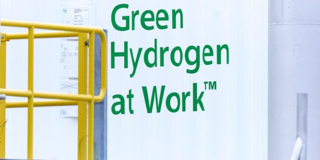 Weißer container mit grüner Aufschrift "Green Hydrogen at Work". Gelbes Geländer im Vordergrund.