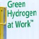 Weißer container mit grüner Aufschrift "Green Hydrogen at Work". Gelbes Geländer im Vordergrund.