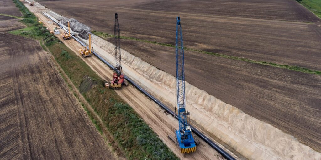 au einer Gasleitung: Ein Seitenbaumleger hebt ein großes Stahlrohr in einen offenen Leitungsgraben, im Hintergrund weitere Baumaschinen und Erdaushub.
