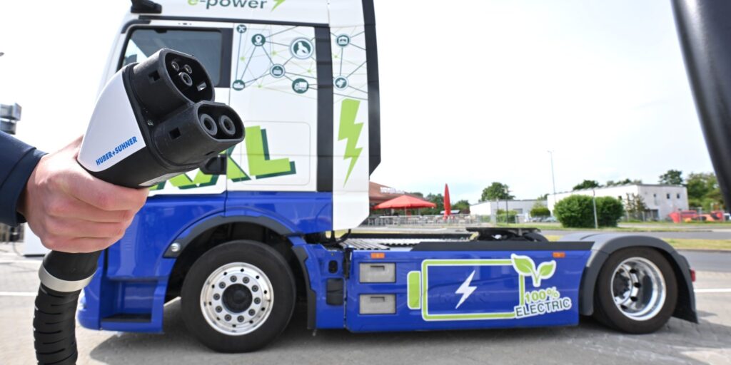 Ladestecker im Vordergrund, dahinter ein blau-weißer Elektro-Lkw mit der Aufschrift „e-power“.