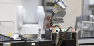 Ein Robotergreifer hält mehrere zylindrische Batteriezellen über einer Halterung, davor steht eine weiße Rundzelle mit Etikett.