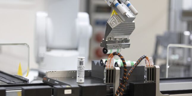 Ein Robotergreifer hält mehrere zylindrische Batteriezellen über einer Halterung, davor steht eine weiße Rundzelle mit Etikett.