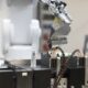 Ein Robotergreifer hält mehrere zylindrische Batteriezellen über einer Halterung, davor steht eine weiße Rundzelle mit Etikett.