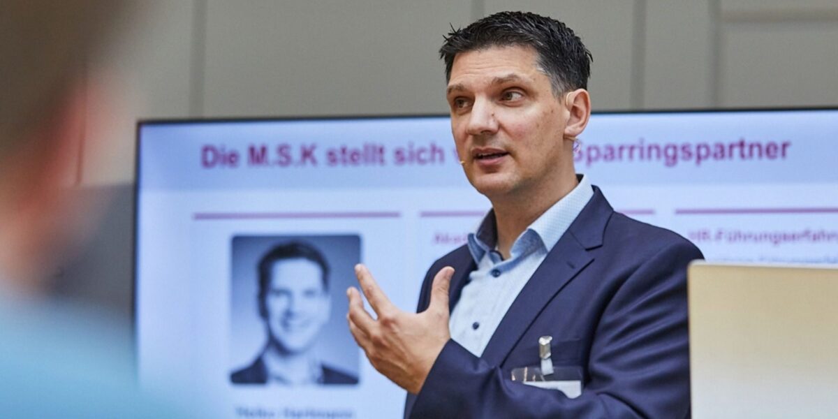 Ein Mann steht vor einem großen Bildschirm und hält einen Vortrag.