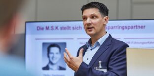Ein Mann steht vor einem großen Bildschirm und hält einen Vortrag.