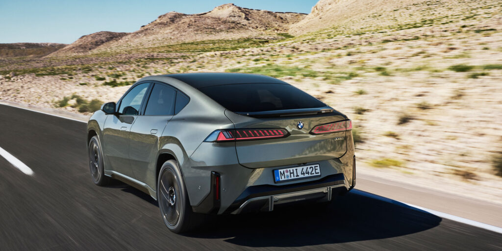 Grauer BMW iX4 fährt auf einer Straße durch eine trockene Landschaft. Die Heckansicht zeigt die coupéförmige Dachlinie, schmale Rückleuchten und den Heckspoiler.