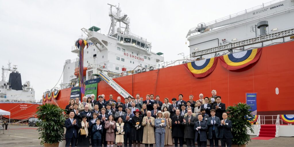Gruppenfoto vor den Gastankern „Antwerpen" und „Arlon" bei der Taufzeremonie auf der HD Hyundai Heavy Industries Werft in Ulsan am 9. April 2026. Im Hintergrund ist der orangefarbene Rumpf eines der beiden Schiffe zu sehen, davor ein Banner mit der Aufschrift „Naming of Antwerpen & Arlon".