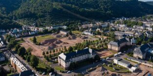 Luftaufnahme der ehemaligen Jägerkaserne in Trier-West mit freigeräumten Bauflächen, Materialhaufen und erhaltenen Kasernengebäuden vor bewaldeten Hängen.