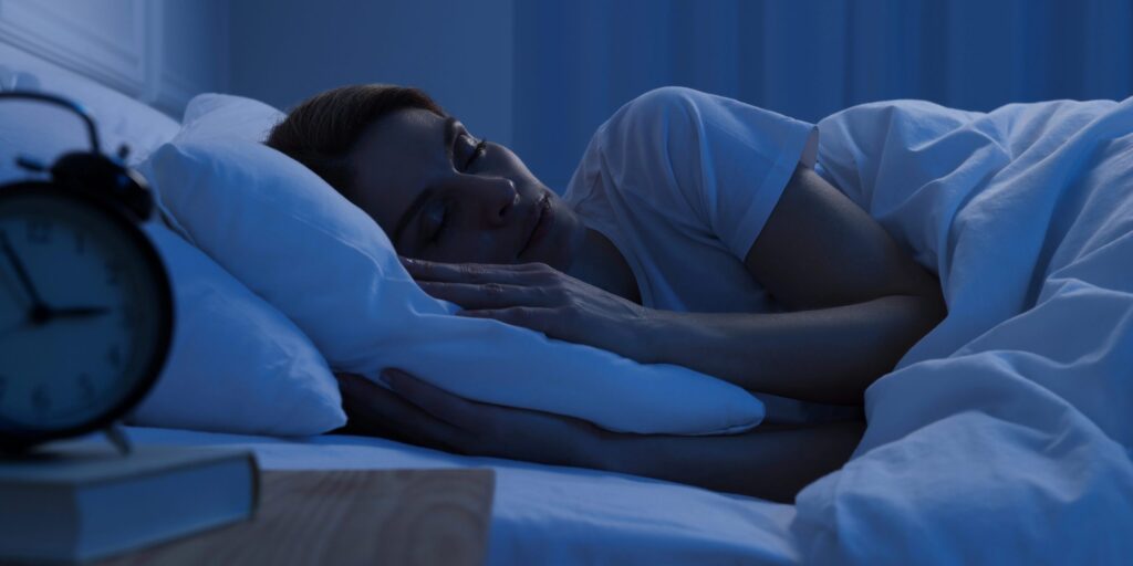 Eine Person schläft nachts seitlich im Bett, der Kopf liegt auf einem Kissen, das Gesicht ist entspannt. Das Zimmer ist dunkel und in blaues Licht getaucht. Im Vordergrund steht ein analoger Wecker auf einem Nachttisch, daneben ein Buch. Die Szene vermittelt ruhigen, überwachten Nachtschlaf.