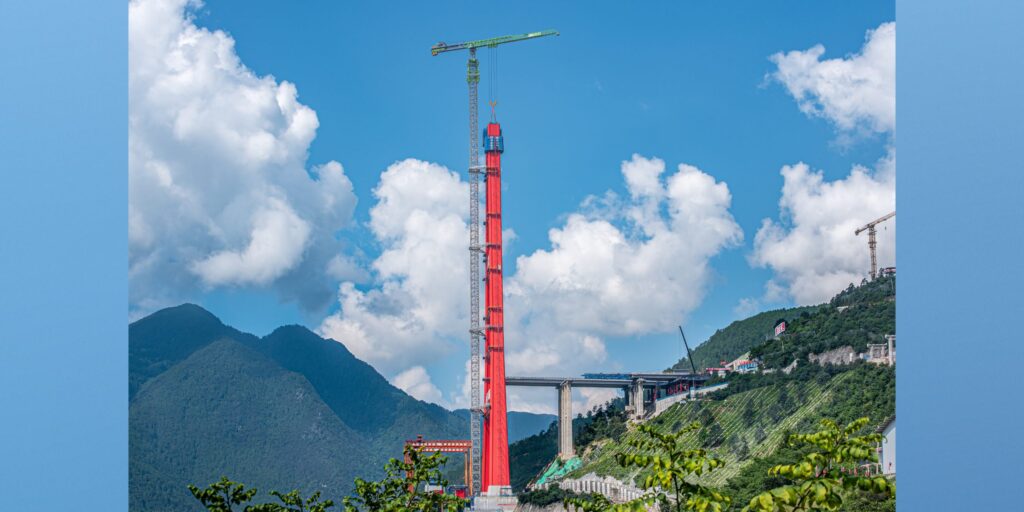 Hoher roter Brückenpylon mit grünem Turmdrehkran auf einer Baustelle im chinesischen Gebirge. Im Hintergrund sind Berge, eine Zufahrtsbrücke und weitere Baukrane zu sehen.