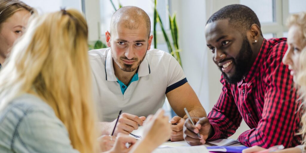 Junge Erwachsene sitzen gemeinsam an einem Tisch, diskutieren und arbeiten konzentriert an Unterlagen – Teamarbeit und Ideenaustausch in einer Lernumgebung.