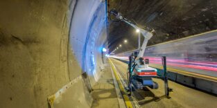 Fischer-Bohrroboter mit ausgefahrenem Arm am Tunnelgewölbe; daneben Lichtspuren vorbeifahrender Fahrzeuge hinter einer Schutzbarriere.