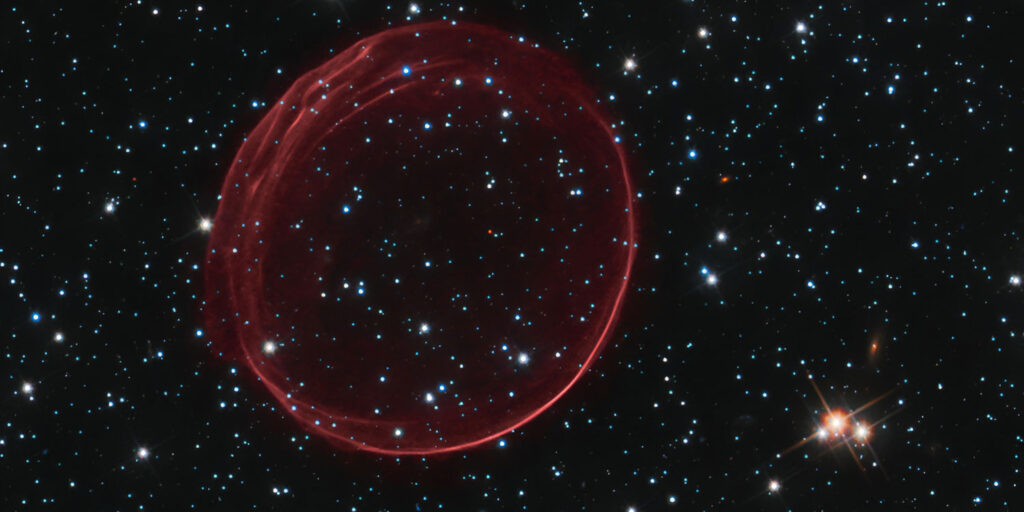 Hubble-Aufnahme des Supernova-Überrests SNR 0509-67.5. Eine große, nahezu kreisförmige rote Gashülle leuchtet vor einem dunklen Sternenhimmel mit zahlreichen hellen Sternen.