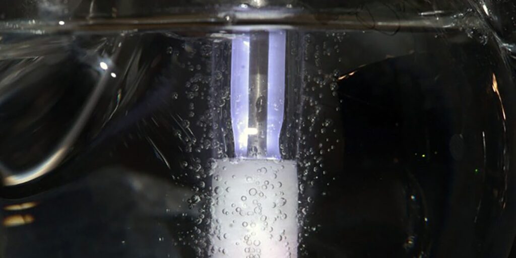Glasrohr im Wasserbad mit einem weiß schimmernden Katalysator-Block und einem violett leuchtenden Plasmabereich