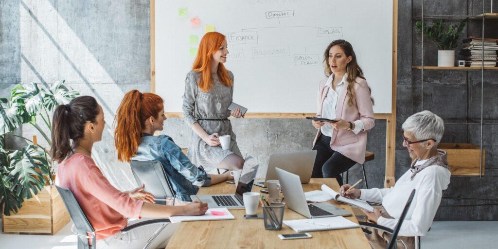 Mehrere Frauen unterschiedlicher Altersgruppen sitzen in einem modernen Besprechungsraum an einem Konferenztisch und diskutieren. Zwei Frauen stehen vor einem Whiteboard mit einer skizzierten Organisationsstruktur und präsentieren Inhalte, während andere Notizen machen oder an Laptops arbeiten. Die Szene vermittelt Teamarbeit, Austausch und Weiterbildung im beruflichen Kontext.