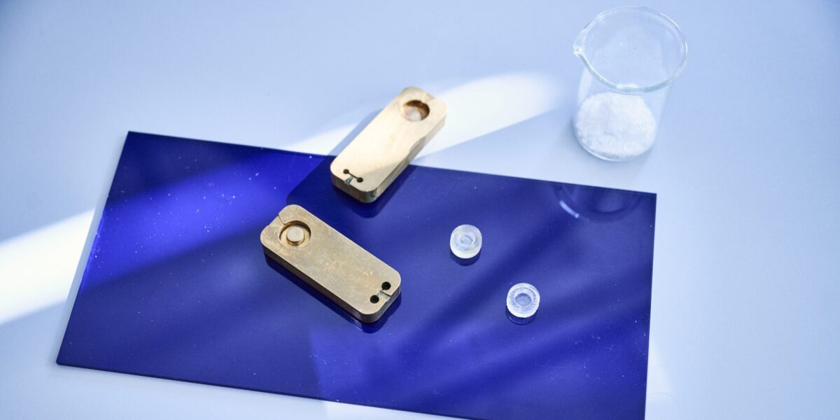 Zwei transparente Mini-Petrischalen neben einem geöffneten metallischen Formwerkzeug auf einer blauen Unterlage; dahinter ein Becherglas mit weißem Material.