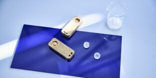 Zwei transparente Mini-Petrischalen neben einem geöffneten metallischen Formwerkzeug auf einer blauen Unterlage; dahinter ein Becherglas mit weißem Material.