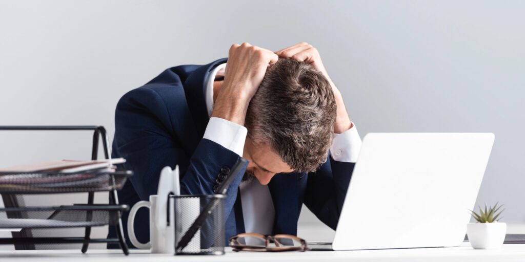 Müder Geschäftsmann sitzt im Büro neben Laptop und Dokumenten im unscharfen Vordergrund.