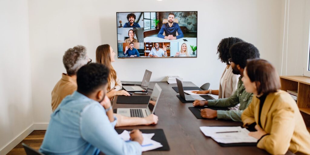 Eine Gruppe von Kollegen sitzt im Konferenzraum, weitere Personen sind digital auf einem Bildschirm zugeschaltet.