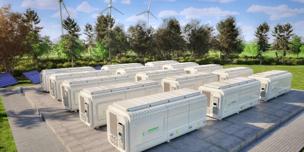 Weiße Batteriespeicher-Container auf einer gepflasterten Fläche, im Hintergrund Bäume, Solarmodule und Windräder unter blauem Himmel.